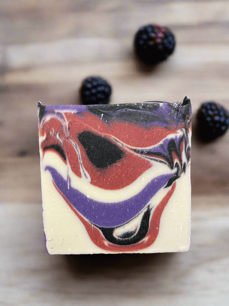 Vegan Black Raspberry Vanilla soap with marbled design, surrounded by fresh blackberries on a wooden surface.