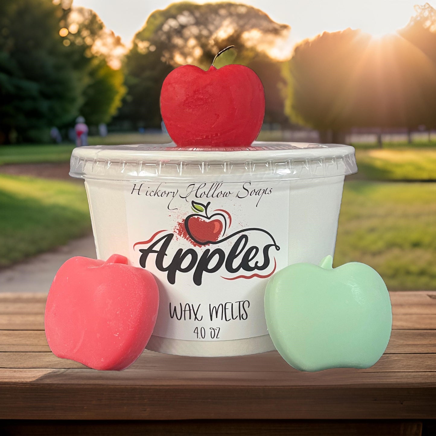 Red and green apple wax melts next to a container labeled 'Apples Wax Melts' in a scenic outdoor setting.