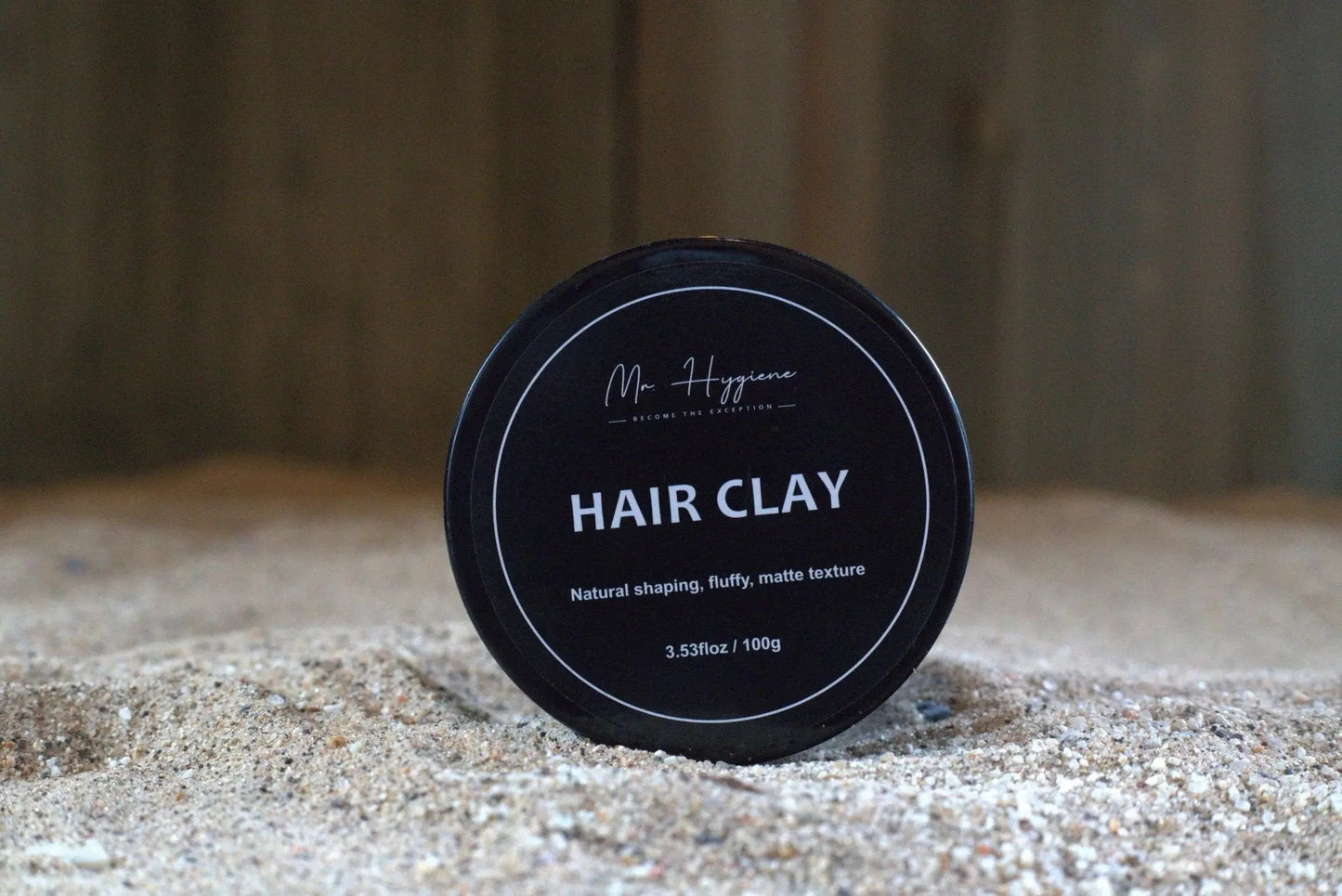 Matte Finish Hair Clay in a black container on sand, providing a strong hold and natural look for styled hair.